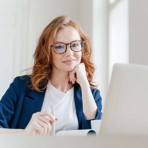 Horizontal shot of pleasant looking successful professional female lawyer learns clients case, works on modern laptop computer, dressed in formal apparel and transparent glasses, poses in office